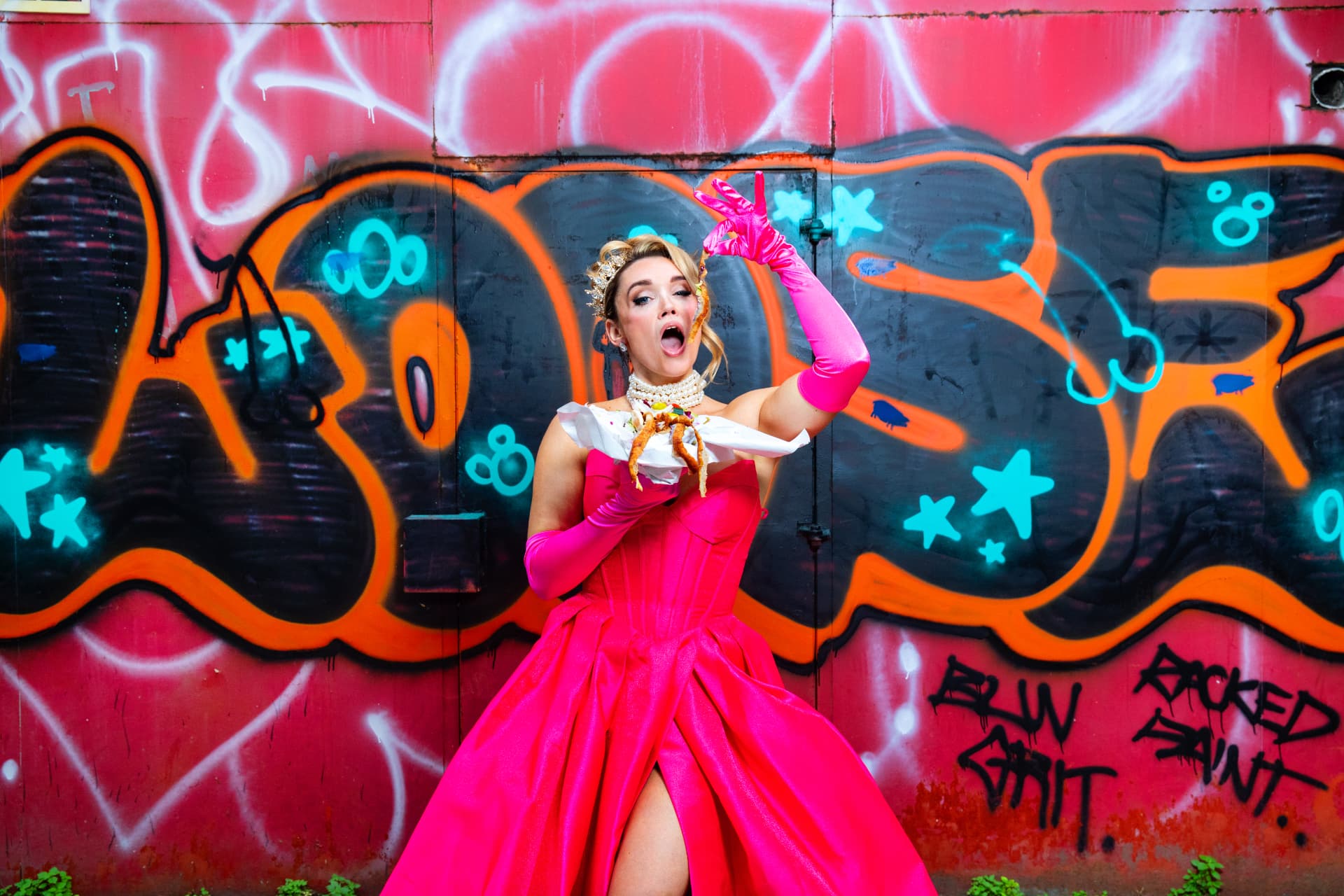 Sophie McCartney in a pink gown and gloves, eating chips in front of graffiti